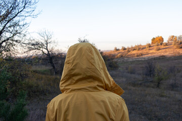 Fototapeta na wymiar A woman in a yellow raincoat with a hood walks through the valley. Back view. Autumn evening landscape