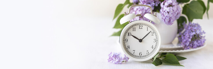Spring change, Daylight Saving Time concept. White alarm clock and lilac flowers on the wooden...