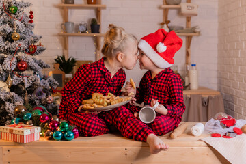children in red pajamas and santa hats and their dog dachshund eat christmas cookies in the kitchen