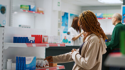 Happy african american client checking boxes of medication on pharmacy shelves, looking at healthcare products to buy prescription treatment. Woman buying medicaments and vitamins.