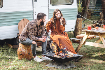 Young beautiful loving couple travelling across country in the van. Millennial man and woman in a travel camper. Cozy atmosphere, vacations vibe. Drinking tea, cooking marshmallow on a campfire