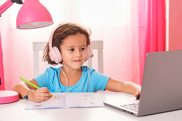 curly brown-haired girl with pink music headphones and a blue t-shirt doing homework with the laptop and a notebook on her desk