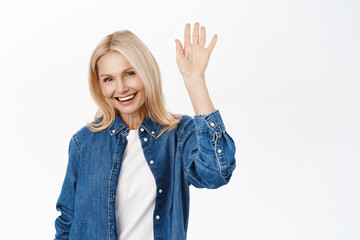 Close up of beautiful blond senior woman, happy lady saying hello, waving hand to say hi, standing...