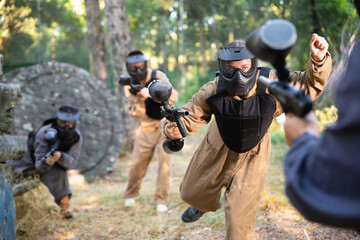 Fototapeta premium Young woman paintball player wearing protective clothes and helmet, rushing through enemy lines, shooting with paintball marker.