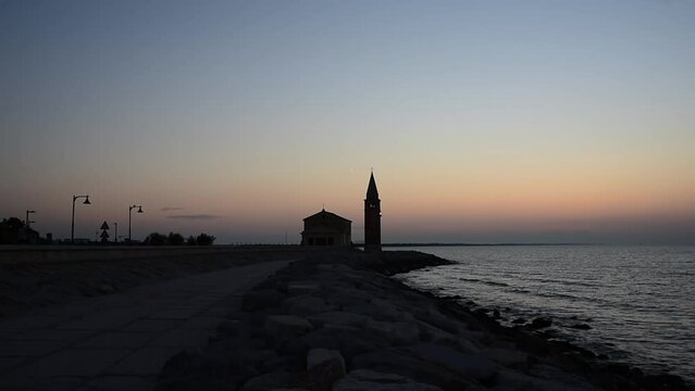 People stroll at dawn on the seafront of Caorle, in the background the sanctuary "Madonna dell'Angelo" built on a promontory surrounded by rocks.
