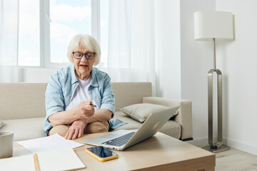 a cheerful elderly woman in stylish home clothes is sitting on a cozy sofa working from home on a laptop and concentrating on auxiliary working tools