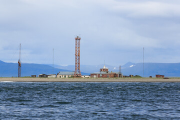 Fototapeta na wymiar Lighthouse by the sea. Lighthouse tower and old buildings and structures on the shore. Marine transport infrastructure in the Far East of Russia. Spit Russkaya Koshka, Chukotka. Anadyr Bay, Bering Sea