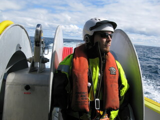 Work boat crew member in high visibility personal protective equipment on small boat back deck. Person wearing SOLAS approved immersion suit and life jacket.