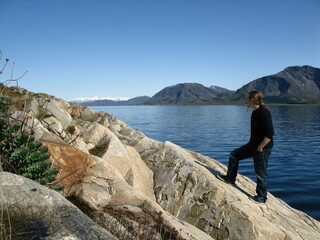 Caucasian bearded long haired male standing on rocky shore line looking towards the fjords and snow capped mountains