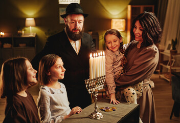 Portrait of modern jewish family looking at silver menorah candle during Hanukkah celebration in cozy home
