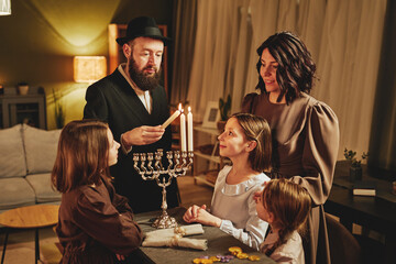 Portrait of orthodox jewish family lighting menorah candle together during Hanukkah celebration - obrazy, fototapety, plakaty