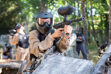 Fototapeta premium Girl in protective paintball outwear standing behind cover, aiming and shooting with paintball marker.