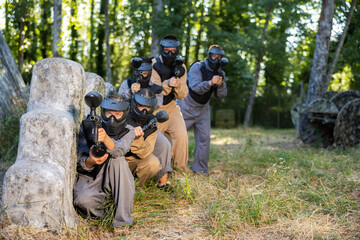 Team taking cover and eliminating opponents on paintball field. People in protective outwear and helmets aiming with paintball markers.