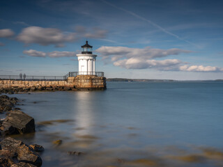 Portland Lighthouse on a bright, sunny fall day. The sky is slightly cloudy. We see a large rock in the foreground. Artistic photo, neutral filter used with a long exposure.