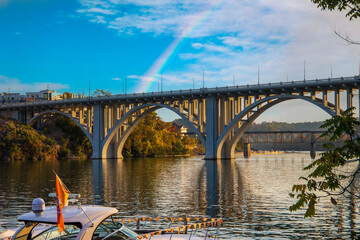 a gorgeous autumn landscape along the Tennessee River at sunset with boats docked on the water and autumn colored trees, lush green trees and the Henley Street Bridge with blue sky and a rainbow - obrazy, fototapety, plakaty