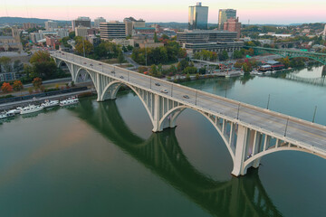 aerial shot of the Henley Street Bridge over the Tennessee River surrounded by autumn colored trees, lush green trees and office buildings with powerful clouds at sunset - obrazy, fototapety, plakaty