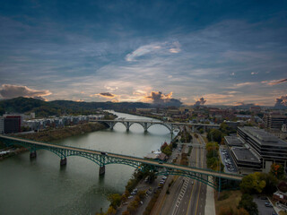 aerial shot of the Henley Street Bridge and Gay Street Bridge over the Tennessee River surrounded by autumn colored trees, lush green trees and office buildings with powerful clouds at sunset - obrazy, fototapety, plakaty