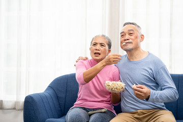 Excited mature couple, Senior man and woman watching tv, senior sport fans celebrating favorite team victory, sitting on cozy couch and eating popcorn snack at home, enjoying weekend. Happy senior.