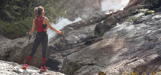 Female trail runner with poles walking standing on mountain top looking at waterfalls.