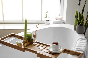 Wooden bathtub tray with candles, eyeglasses and cup of tea in light room