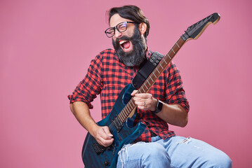 Bearded man playing his electric guitar, singing along, having fun; casual wear, wearing glasses; pink studio background