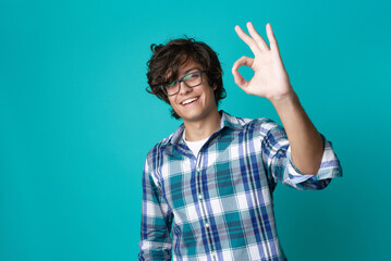 Young man with spectacles making an ok sign, expressing approval and looking cheerful and content;...