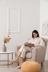 Young woman reading magazine in beige armchair at home