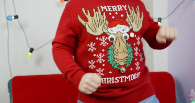 Fat person in knitted Christmas sweater with picture of deer with bell is dancing funny, waving arms and shaking stomach, close up. Garland with colorful light bulbs is decorate room