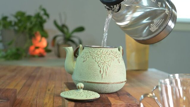 Making herbal tea in a green teapot on a wood table in a light and airy room with green plants in the background and a glass teacup