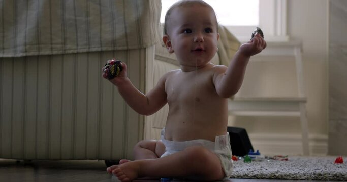 Baby girl sitting and drooling on living room floor with toys in hands - looks at camera then looks away