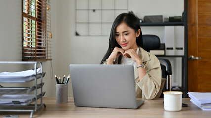 Charming Asian businesswoman sits at her desk, looking at her laptop screen
