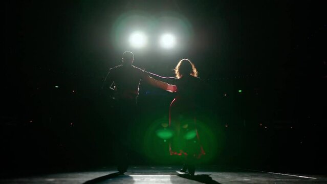 Ballroom dancers slowly coming out on stage holding hands across and swaying hips,under 2 bright spotlights.Rear view,silhouettes wearing dance costumes,red dress.Camera moves back in far distance.