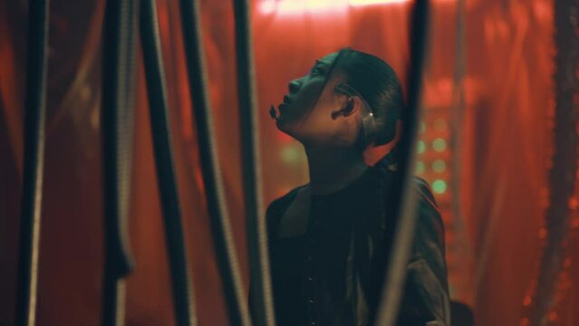 Cyberpunk girl trapped and tries to escape using hand gestures. Asian girl in cyberpunk style wearing headset with LED lights communicates with someone using microphone. Black thick rods hanging.