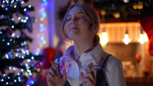 Teenage girl crossing fingers making wish on Christmas night at home indoors. Portrait of charming confident Caucasian teenager wishing miracle on New year's eve in darkness looking away smiling