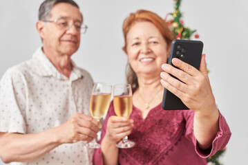 Happy senior hispanic couple talking by video call while toasting during Christmas or New Year, celebrating holidays at home, together. Selective focus on the hand holding the smartphone.