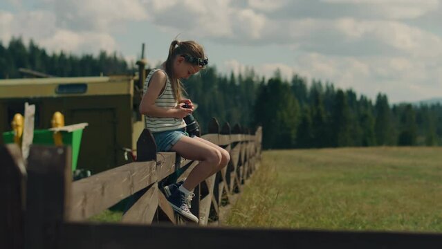 Young photographer changing camera settings. Girl sitting on wooden fence, concentrating on process, shooting beautiful nature landscape, spending leisure time outdoor. Slow motion.