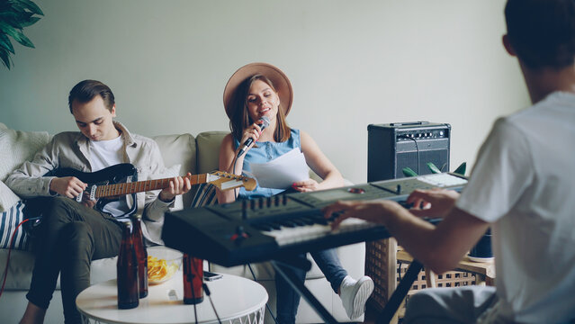 Attractive Young People Musical Band Are Holding Rehearsal In Nice Room At Home, Playing Musical Instruments And Singing In Microphone. Music And Millennials Concept.