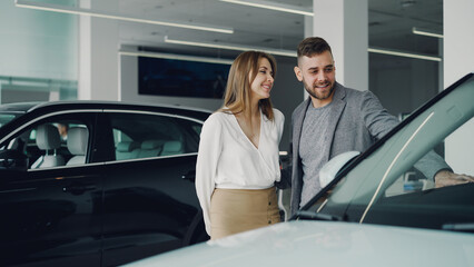 Fototapeta na wymiar Attractive young couple confident bearded guy and his smiling pretty girlfriend are choosing new automobile together looking at luxury car in auto dealership and talking.