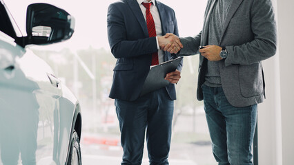 Motor showroom employee is giving car key to young male buyer and shaking hands, dealer is holding documents and gesturing. Selling and buying luxury transport concept.