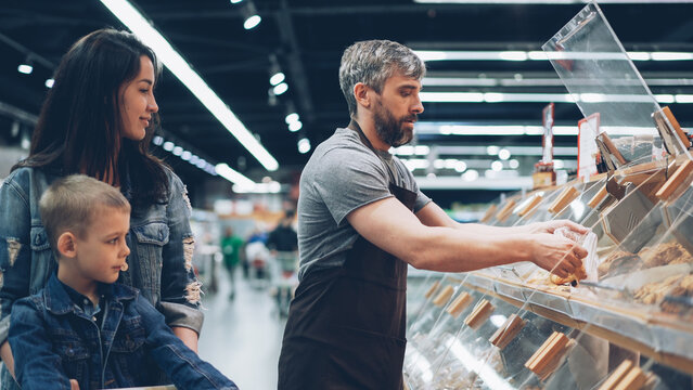 Male shop assistant is working in bakery department in supermarket then packing bread roll for nice family mother and son and giving food to attractive woman. - Powered by Adobe
