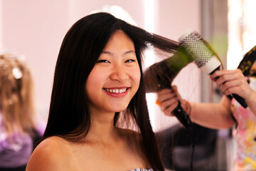 hair stylist combing with brush and dryer the straight hair of a young asian woman in the...