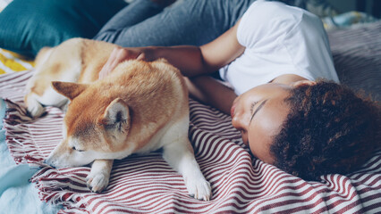Pretty African American teenage girl and her loyal dog are sleeping together on bed at home, person is hugging animal in sleep. Trust, love and rest concept.