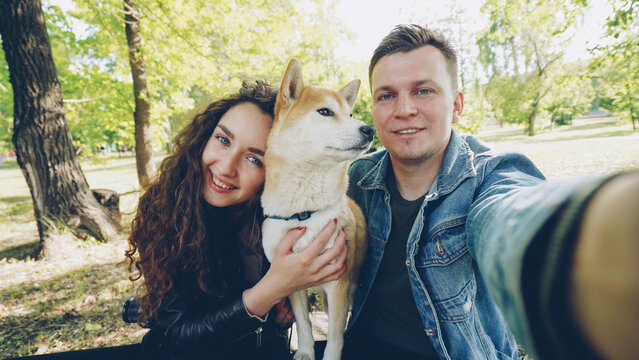 Point of view shot of cheerful married couple taking selfie with pet shiba inu dog, young people are talking, laughing and caressing the animal while looking at camera.