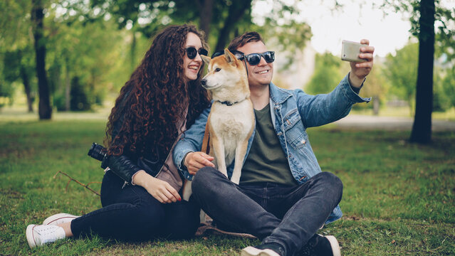 Handsome young man is taking selfie with his pretty wife and cute dog, all wearing sunglasses. Guy is holding smartphone taking funny pictures and posing.