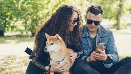Young pretty woman is petting her dog sitting on grass while her boyfriend is showing her smartphone screen, people are talking and laughing. Weekend in the park concept.
