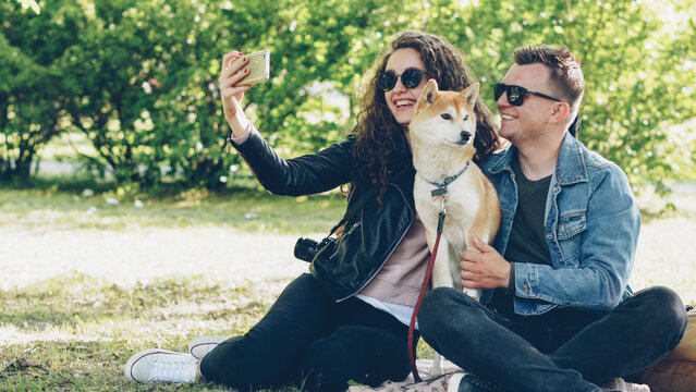 Happy couple is taking selfie with their dog using smartphone sitting on grass in the park, people are posing, talking and petting shiba inu puppy. Photograph and animals concept.