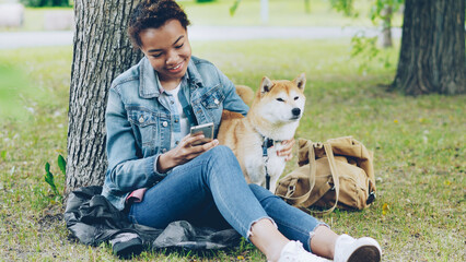 Happy African American woman is using modern smartphone and caressing her cute pet dog resting in city park on windy summer day. Nature, animals and people concept.