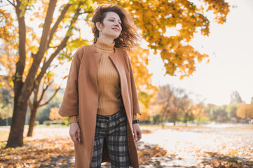 Red-haired middle-aged woman walks in the autumn park and collects yellow maple leaves. Middle-aged woman in an orange coat and a beige sweater on a walk in nature in autumn during leaf fall