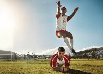 Fototapeta na wymiar Soccer, fitness and training with a sports man jumping over his teammate during practice on a grass pitch or field. Football, workout and exercise with a male athlete exercising with his team outdoor