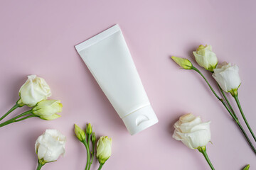 Blank plastic white tube for cream, mask, shampoo or toothpaste on pink background among eustoma flowers. Spring presentation of cosmetics package. Mockup